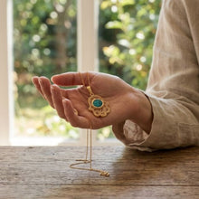 A gold pendant necklace with a turquoise-blue stone is held in a person's hand over a rustic wooden table, with a softly blurred window and greenery in the background.