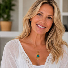 A smiling blonde woman wearing a gold Hamsa hand pendant necklace with a turquoise blue stone, set against a softly blurred indoor background.