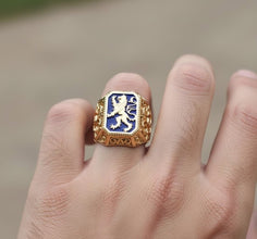 Gold ring with a blue emblem featuring a lion on a finger against a blurred background