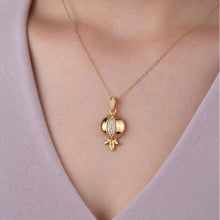 A gold pendant necklace featuring a stylized pomegranate design with a pavé-set clear stone center, shown worn on a model's neck over a light purple V-neck top.