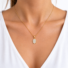 A gold Hamsa hand pendant necklace covered in small clear stones, worn on a model's neck over a white V-neck top.
