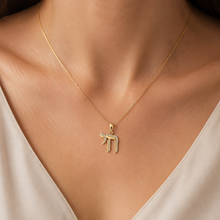 A delicate gold chain necklace with a Hebrew letter pendant, worn by a model in a close-up shot. The pendant is adorned with small clear stones, and the model is wearing a light beige satin top.