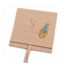 A gold Hamsa hand pendant with a blue cabochon gemstone, displayed on a beige suede jewelry pouch branded with 'YOELS JEWELRY'.