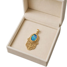 A gold hamsa hand pendant with a blue opal inlay, presented inside an open beige jewelry box on a white background.