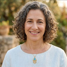 A smiling woman with curly gray-brown hair wearing a light blue top and a gold pendant necklace with a blue stone, photographed outdoors against a softly blurred garden background.