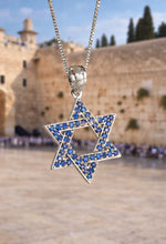 Star of David pendant with blue stones on a chain, set against a blurred background of a stone wall and trees.