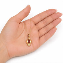 A gold pendant necklace shaped like a pomegranate with red gemstone accents, displayed on an open hand against a white background.