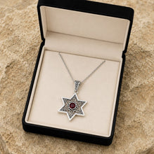 Silver star-shaped pendant with a red gemstone in a black jewelry box on a stone surface