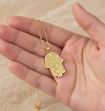 Gold Hamsa hand necklace held in a hand against a beige background