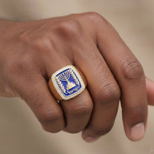 Hand wearing a ring with a blue and gold emblem on a neutral background