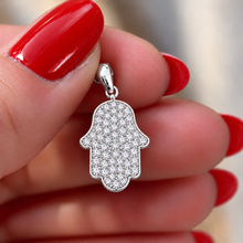A gold Hamsa hand pendant covered in small clear stones, held between fingers with dark red manicured nails.