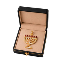 Gold menorah pendant with red stones in a black box on a beige background