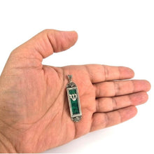 A silver-toned rectangular pendant with a green inlay and a white crescent moon symbol, shown resting on an open hand against a white background.