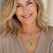 A smiling blonde woman wearing a gold Hamsa hand pendant necklace with a turquoise blue stone, set against a softly blurred indoor background.