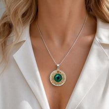 A close-up of a woman wearing a silver chain necklace with a round pendant featuring a green center and gold-toned textured detailing, styled with a white satin blouse.