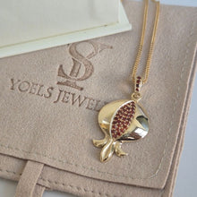 A gold pendant necklace shaped like a pomegranate with a red crystal-studded seed, displayed on a beige suede jewelry pouch embossed with 'YOELS JEWELRY'.