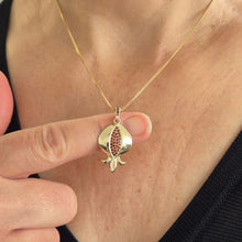 A gold pendant necklace featuring a stylized pomegranate design with a red textured center, worn on a model's neck and held closer by a hand.