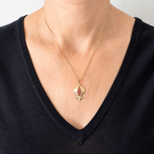 A gold pendant necklace featuring a stylized pomegranate with red and gold enamel detailing, shown worn on a model's neck over a black V-neck top.
