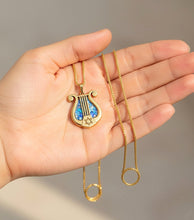 Gold necklace with a lyre-shaped pendant held in a hand against a neutral background