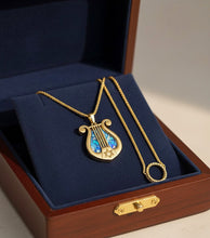Gold necklace with a lyre-shaped pendant on a blue cushion in a wooden box.