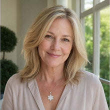 A smiling blonde woman wearing a gold Star of David pendant necklace and a light beige blouse, photographed in a bright indoor setting with a window and greenery in the background.