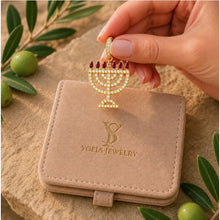 Gold Menorah pendant with red stones held above a Yoel's Jewelry box on a stone surface with green leaves.