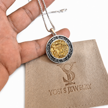 A silver and gold pendant necklace held in a hand, featuring a circular coin-style pendant with Hebrew text and a central gold emblem, displayed above a beige Yosi's Jewelry pouch on a white background.