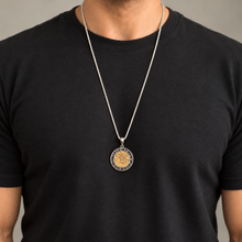 A silver chain necklace with a circular pendant featuring a gold-toned central design and a black outer ring, worn by a person in a black t-shirt against a neutral gray background.