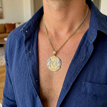 A close-up of a man wearing a gold chain necklace with a circular pendant featuring a lion and unicorn design, paired with an open blue button-down shirt.