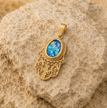 A gold Hamsa hand pendant with an oval blue opal center, resting on a textured beige rock.