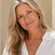 A woman with blonde hair wearing a gold Hamsa hand pendant necklace with a blue stone, dressed in a white collared shirt against a neutral beige background.