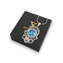 A silver Hamsa hand pendant necklace with a blue and green iridescent stone in the center, displayed on a black gift box with gold lettering.