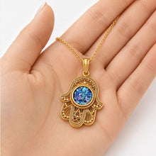 A gold Hamsa hand pendant necklace with a blue mosaic center, held in the palm of a hand against a clean white background.