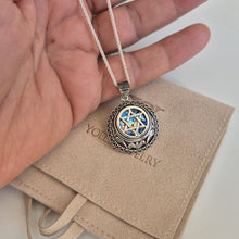 A silver-toned pendant necklace with a Star of David and blue enamel details, held in a hand over a beige jewelry pouch embossed with 'YOUR JEWELRY'.