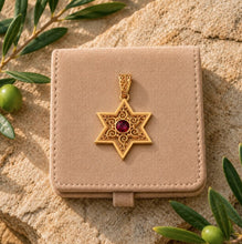 Gold star-shaped pendant with a red gemstone on a beige velvet box, surrounded by green leaves.