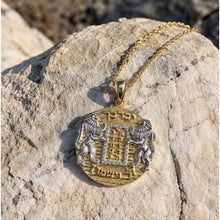 A gold and silver pendant necklace displayed on a textured light gray rock outdoors. The pendant features Hebrew text and a central Hebrew cross design, with two lions on either side.